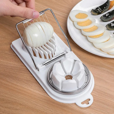 Multifunctional stainless steel egg slicer and splitter tool on wooden surface; sliced eggs on plate in background.